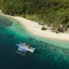 Aerial view of a blue outrigger boat anchored in turquoise water beside a white sand beach lined with palm trees